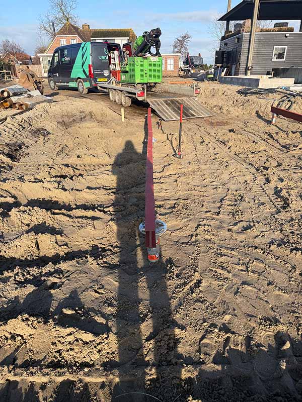 fundering-mantelzorg-woning Close-up van een rode schroefpaal die in de grond wordt geschroefd door een boorinstallatie van Van Ham Funderingstechnieken, zichtbaar op een aanhanger op de achtergrond.