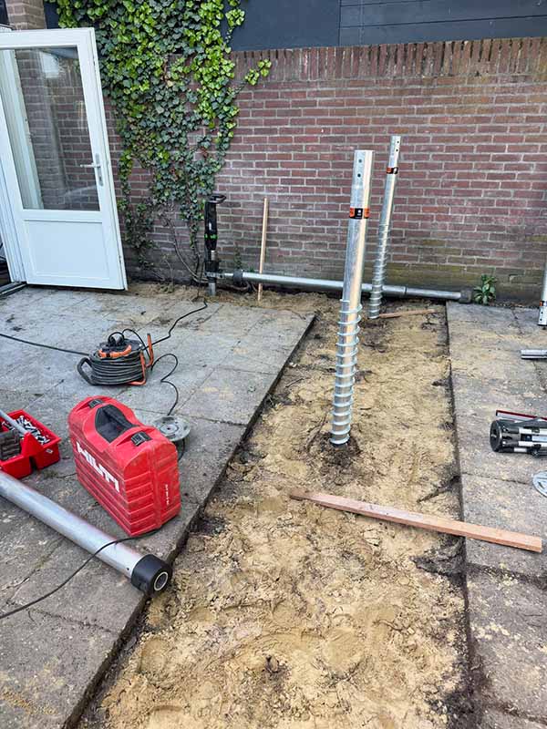 Twee geplaatste V76 schroefpalen steken uit de zandbodem langs de achtergevel, klaar als fundering voor de aanbouw van een hoekwoning in 's-Hertogenbosch